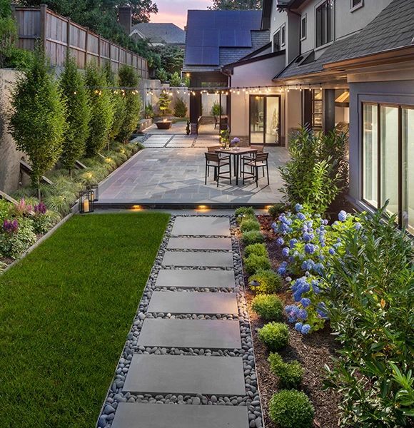 Modern backyard with a stone pathway flanked by manicured grass and vibrant blue hydrangeas. A patio with string lights and seating creates a cozy ambiance.