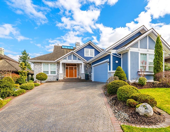 A charming suburban house with a stone facade and blue siding. It features a neatly paved driveway, manicured shrubs, and a bright blue sky.