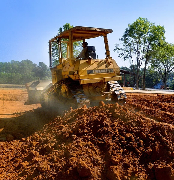 A yellow bulldozer moves dirt on a construction site, stirring dust under a clear blue sky. Trees in the background add a natural contrast.