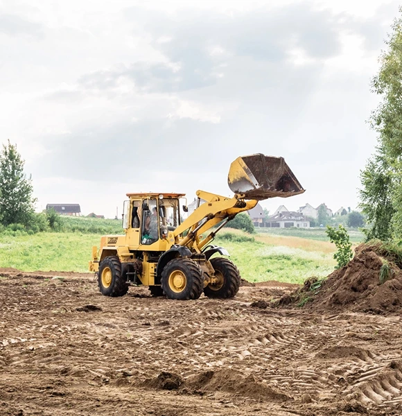 A yellow bulldozer with a raised bucket works on a large, muddy field. Green fields and distant houses are visible under a cloudy sky.