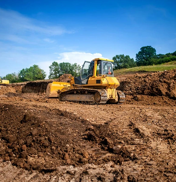 A yellow bulldozer moves earth on a construction site under a clear blue sky. Surrounding green trees contrast with the rich brown soil, evoking a sense of progress.