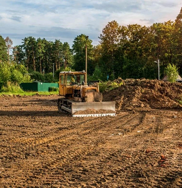 A bulldozer is parked on a dirt construction site surrounded by trees under a cloudy sky. The scene conveys a sense of land clearing progress and nature.