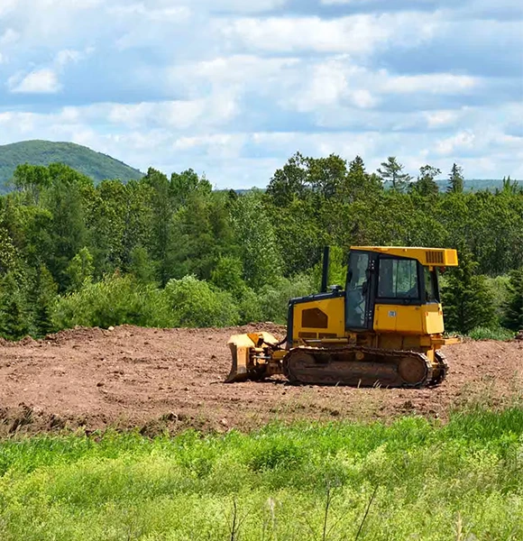 A yellow bulldozer sits on a freshly cleared dirt area, surrounded by lush green trees and hills under a partly cloudy blue sky, conveying calm industry.