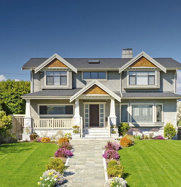 Two-story house with a gray facade, triangular roof accents, and a neat front lawn. Vibrant flowers line the pathway, under a clear blue sky.