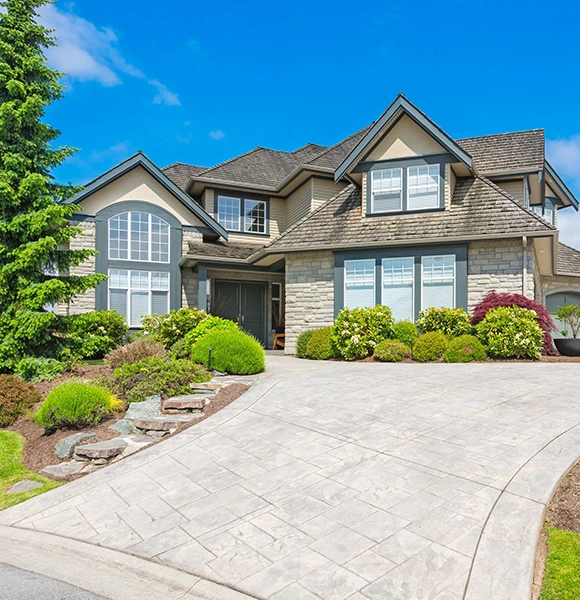 Elegant suburban house with stone facade, large windows, and gabled roof under a clear blue sky. Lush gardens and curved driveway enhance curb appeal.