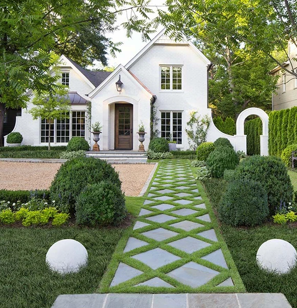 Charming white cottage with dormer windows, a wooden door, and lush green landscaping. A geometric stone path leads through a manicured garden, evoking elegance.